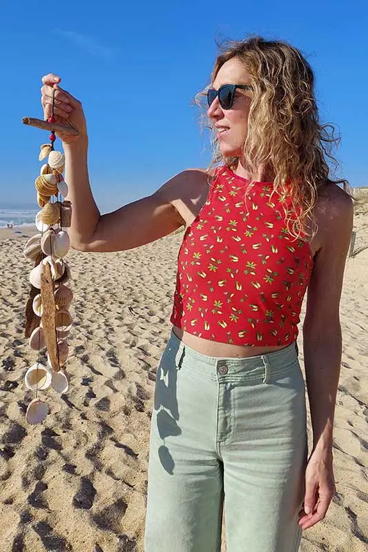 Primer plano de Ana Nushu en la playa, sonriendo mientras sostiene un móvil decorativo vertical hecho con conchas naturales y madera varada.