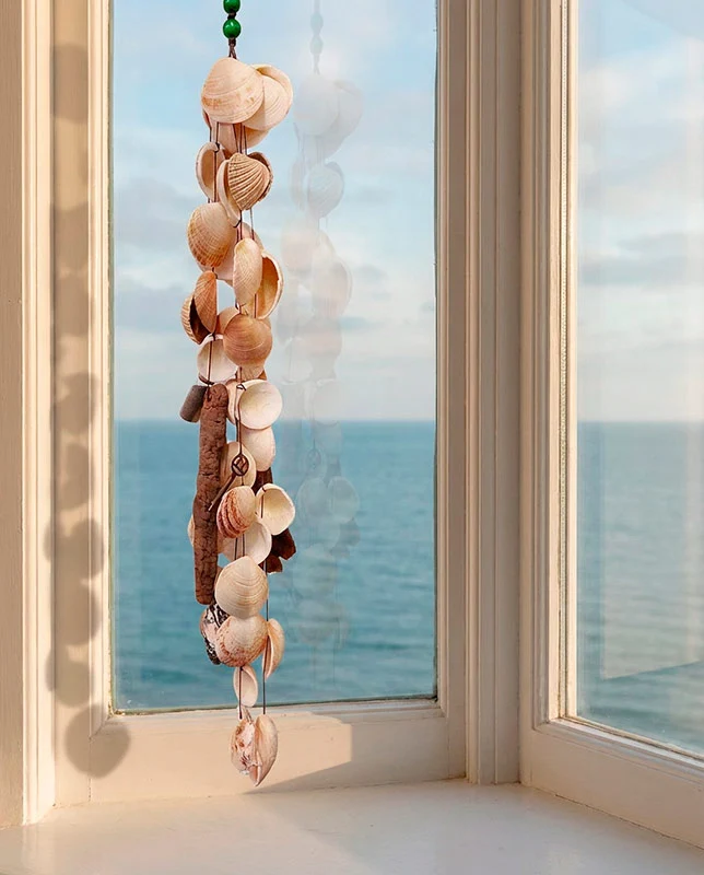conchas-naturales-colgante Colgante de techo rcon conchas marinas y un trozo de madera varada, suspendido frente a una ventana con vistas al mar azul.