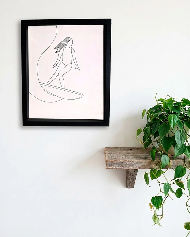 Lámina artística con un dibujo lineal minimalista de una mujer surfeando, colgada en una pared blanca.